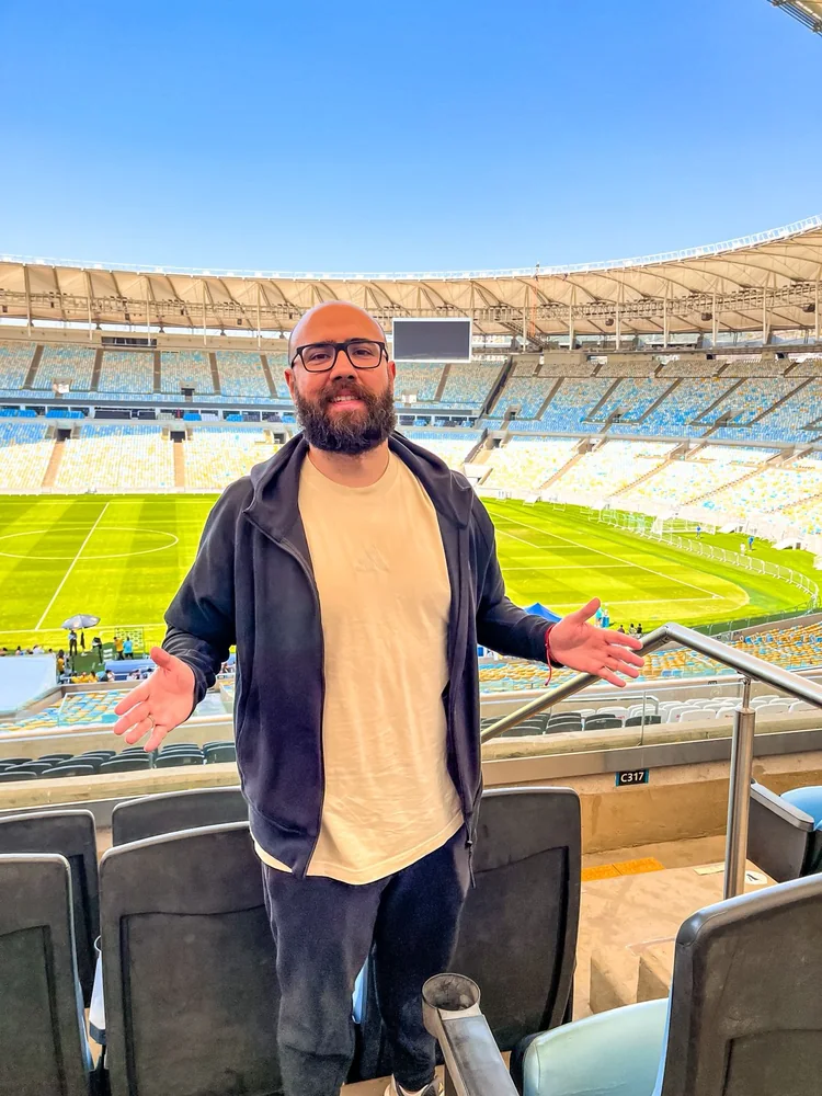 Digão com os braços abertos em um estádio, com o campo e a arquibancada ao fundo