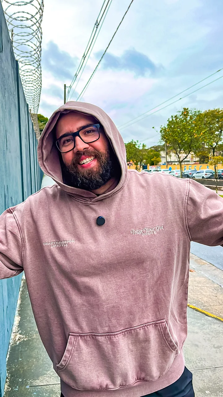 Digão sorrindo, de óculos e moletom com capuz, em uma rua