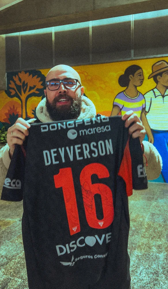 Digão segurando uma camisa autografada para a câmera, em um corredor interno com mural amarelo ao fundo